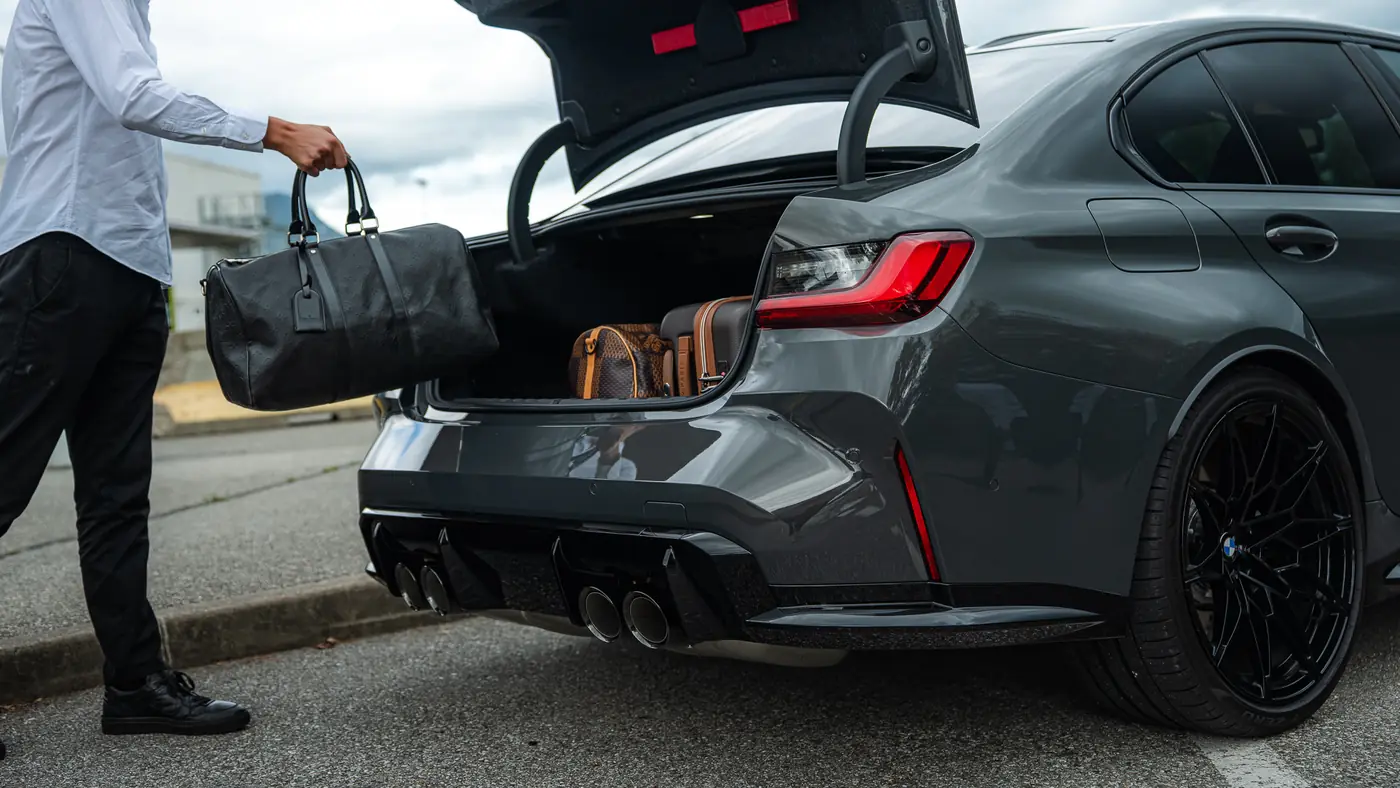 Bagages chargés dans le coffre d'une BMW M3 Compétition Corsiva, sur le parking d'un aéroport