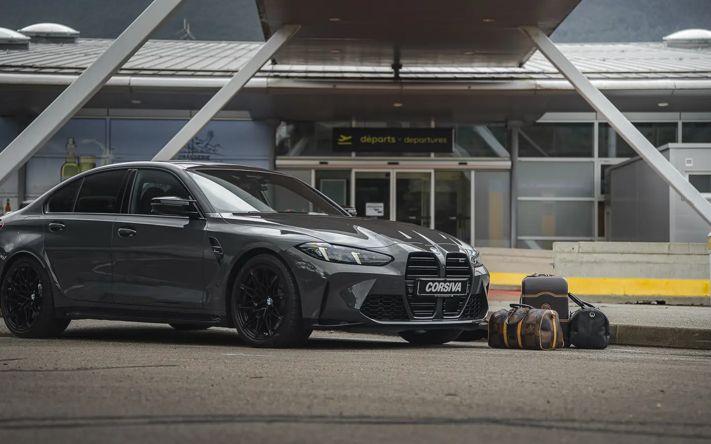 BMW M3 Compétition Corsiva devant un terminal d'aéroport, bagages posés à côté