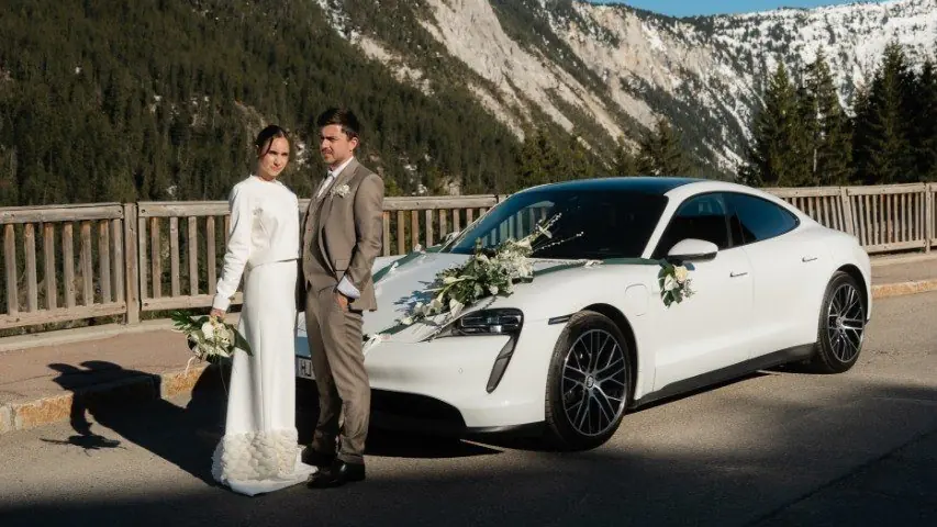 Jeunes mariés devant une Porsche Taycan Corsiva décorée, en montagne