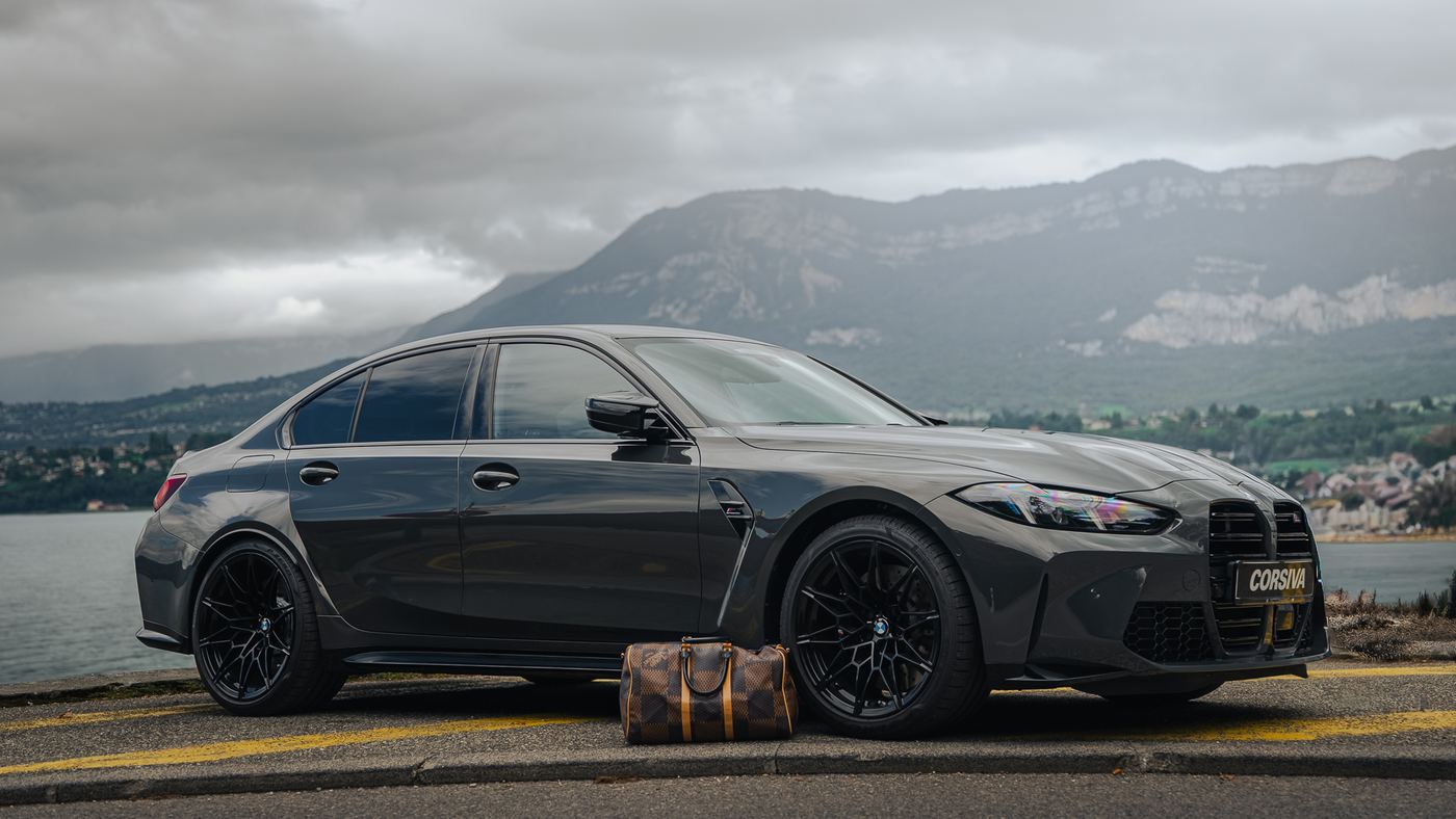 BMW M3 Compétition Corsiva au bord d'un lac alpin, un sac de voyage posé devant la roue